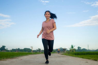 Açık havada koşu antrenmanı. Genç, mutlu ve kendini işine adamış Asyalı bir kadın sağlıklı yaşam tarzında spor ve kardiyo aktivitelerinin keyfini çıkarmak için mavi gökyüzünün altında güzel bir kırsal yolda koşuyor.