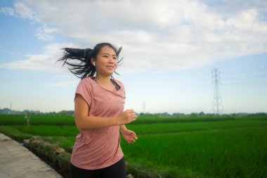 Açık havada koşu antrenmanı. Genç, mutlu ve kendini işine adamış Asyalı bir kadın sağlıklı yaşam tarzında spor ve kardiyo aktivitelerinin keyfini çıkarmak için mavi gökyüzünün altında güzel bir kırsal yolda koşuyor.