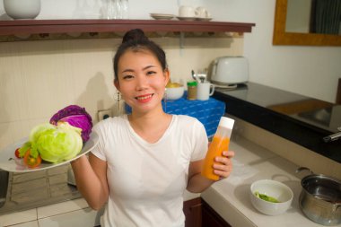 Genç, güzel ve mutlu Asyalı bir kadının portresi. Mutfakta rahat bir şekilde sebze salatası hazırlıyor. Sağlıklı beslenme ve yemek pişirme konseptiyle gülümsüyor.