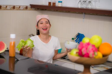 Genç, güzel ve mutlu Asyalı bir kadının portresi. Mutfakta rahat bir şekilde sebze hazırlıyor. Sağlıklı beslenme ve yemek pişirme konseptiyle gülümsüyor.