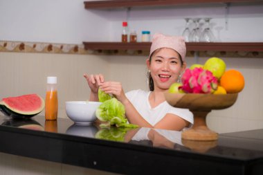Genç, güzel ve mutlu Asyalı bir kadının portresi. Mutfakta rahat bir şekilde sebze hazırlıyor. Sağlıklı beslenme ve yemek pişirme konseptiyle gülümsüyor.
