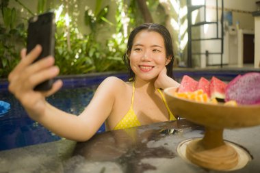 Güzel ve mutlu Asyalı kadın bikinili, villa ya da otel havuzunda cep telefonuyla selfie çekiyor güzel ve rahatlatıcı tatil yerlerinde lüks yaz tatillerinin tadını çıkarıyor. 
