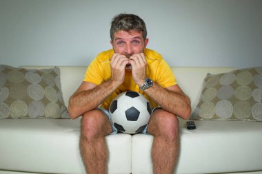 Genç heyecanlı ve gergin futbol taraftarı televizyonda maç seyreden adam oturma odasındaki kanepede tırnaklarını yiyerek yoğun bir duygu ve stres ifadesi sergiliyor.