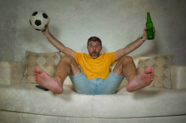 Genç, heyecanlı ve mutlu futbol taraftarı. Oturma odasındaki kanepede elinde bira şişesiyle futbol maçı izleyen adam. Takımın gol atmasını kutluyor.