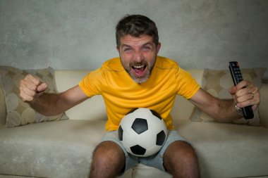 Genç, heyecanlı ve mutlu futbol taraftarı. Oturma odasındaki kanepede futbol maçı seyrediyor. Elinde uzaktan kumandalı televizyon var.