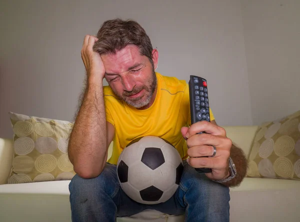 frustrated football fan in intense emotion - home portrait of young dejected and sad man watching soccer game on television at living room couch his team losing the match