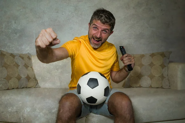 young excited and crazy happy football supporter man watching soccer game on television at living room sofa holding TV remote celebrating team scoring goal feeling intense emotion