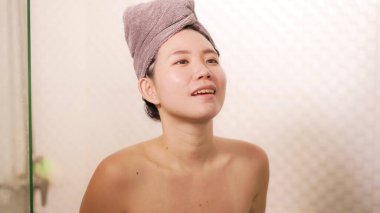 Havluya sarılmış güzel ve mutlu Asyalı bir kadının ev yapımı portresi. Havluya sarılmış, sabah duşundan sonra taze ve neşeli gülümsüyor. Sabah rutini ve hijyeninin keyfini çıkarıyor.