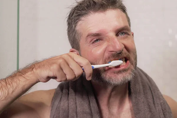 home lifestyle portrait of young attractive and happy man with towel on his neck brushing his teeth in the bathroom relaxed and cheerful enjoying dental care routine