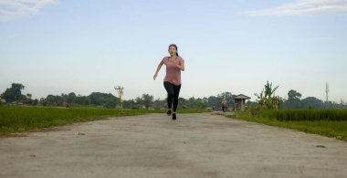 Açık havada koşu. Genç, mutlu ve kendini işine adamış Asyalı bir kadın sağlıklı yaşam tarzındaki spor ve kardiyo aktivitelerinin keyfini çıkarmak için mavi gökyüzünün altında güzel bir kır yolunda egzersiz yapıyor.