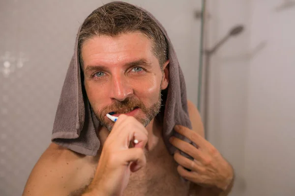 home lifestyle portrait of young attractive and happy man with towel on his head brushing his teeth in the bathroom relaxed and cheerful enjoying dental care routine