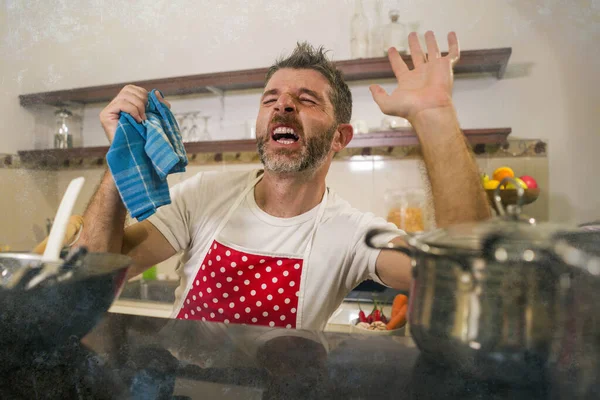 domestic chores stress - young attractive overworked and depressed man in red apron at home kitchen feeling stressed and tired of cooking and dishwashing in frustrated face expression