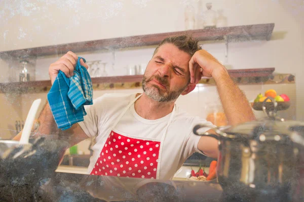 domestic chores stress - young attractive overworked and depressed man in red apron at home kitchen feeling stressed and tired of cooking and dishwashing in frustrated face expression
