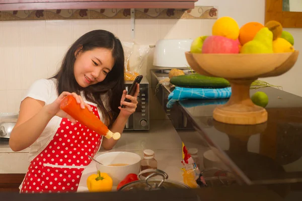 young happy and beautiful Asian Korean home cook woman in red apron reading healthy recipe in mobile phone cooking cheerful preparing delicious meal in kitchen