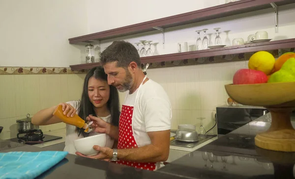 young beautiful and happy mixed ethnicity couple in love cooking together at home kitchen the caucasian husband in red apron and his Asian wife helping him and enjoying cheerful