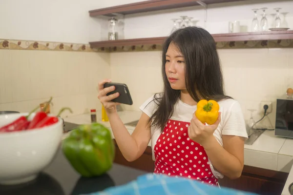 young happy and beautiful Asian Korean home cook woman in red apron reading healthy recipe in mobile phone cooking cheerful preparing delicious meal in kitchen