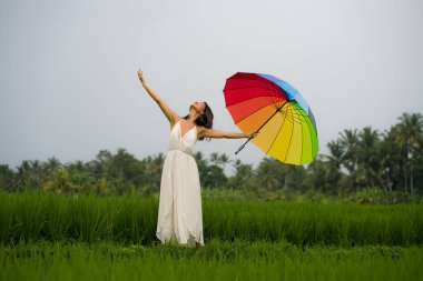 Açık havada genç mutlu ve güzel Asyalı kadın portresi beyaz elbise ve gökkuşağı şemsiyesi içinde doğa kaygısızca geziyor. 