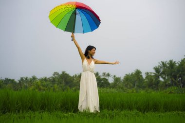 Açık havada, beyaz elbiseli ve gökkuşağı şemsiyeli mutlu ve güzel Asyalı bir kadının tropik yeşil arazide tasasız bir şekilde dolaşmasının resmi. 