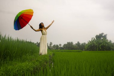 Açık havada, beyaz elbiseli ve gökkuşağı şemsiyeli genç mutlu ve güzel Asyalı bir Japon kadının tropik yeşil arazide tasasız bir şekilde dolaşmasının resmi. 