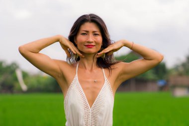 Açık havada tatil portresi. Çekici ve mutlu orta yaşlı Asyalı beyaz elbiseli kadının yeşil alan manzarasında özgürlük ve doğanın tadını çıkarırken tasasız ve neşeli.