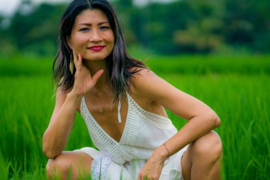 Açık havada tatil portresi. Çekici ve mutlu orta yaşlı Asyalı beyaz elbiseli kadın. Yeşil alan manzarasında özgürlüğün ve doğanın tadını çıkartıyor.