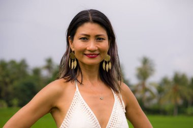 Açık havada tatil portresi. Çekici ve mutlu orta yaşlı Asyalı beyaz elbiseli kadının yeşil alan manzarasında özgürlük ve doğanın tadını çıkarırken tasasız ve neşeli.