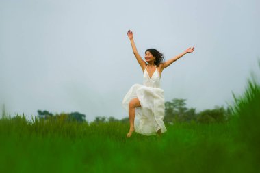 Açık havada tatil portresi, beyaz elbiseli çekici ve zarif Asyalı kadının mutlu ve özgür bir şekilde zıplaması tropikal yeşil alanda doğanın güzelliğinin tadını çıkarması neşeli ve kaygısız