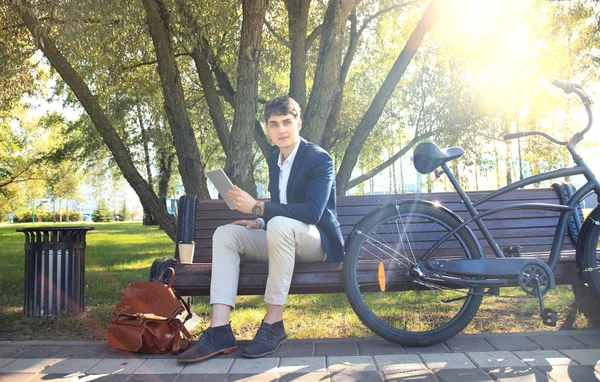 Uomo d'affari in pausa caffe '. È seduto su un banco e lavora al touchpad, accanto alla bici . — Foto Stock