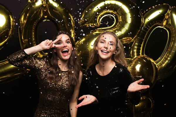 Two happy girls in shiny dresses posing while standing with gold colored 2020 number balloons on black background. — Stock Photo, Image