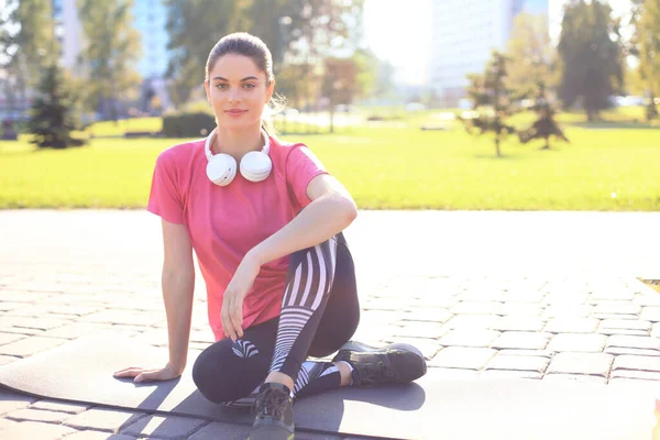 Mulher bonita olhando para longe enquanto sentada no tapete de exercícios no parque, relaxando após o treinamento . — Fotografia de Stock