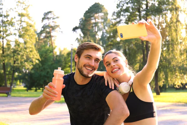 Młoda sportowa para w odzieży sportowej siedzi na parapecie w miejskim parku i robi selfie po treningu w letni słoneczny dzień. — Zdjęcie stockowe