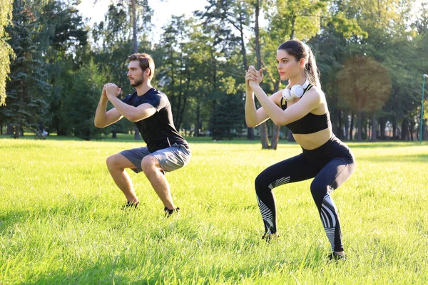 Fiatal sportos férfi és nő együtt edzenek és guggolnak a zöld parkban a nyári napsütéses napokon. — Stock Fotó
