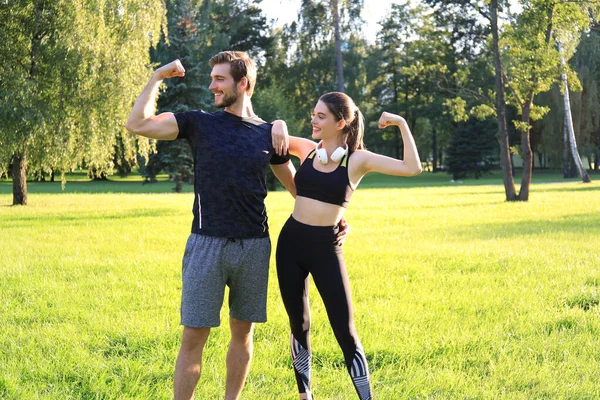 Alegre casal esportivo treinando juntos e mostrando bíceps no parque verde no dia ensolarado . — Fotografia de Stock