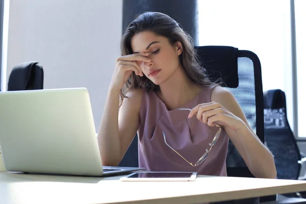 Frustrierte Geschäftsfrau wirkt erschöpft, während sie an ihrem Arbeitsplatz sitzt und ihre Brille in der Hand hält — Stockfoto