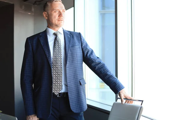 Зрелый бизнесмен в корпоративном костюме, стоящий в офисе и с оптимизмом смотрящий в большие окна . — стоковое фото