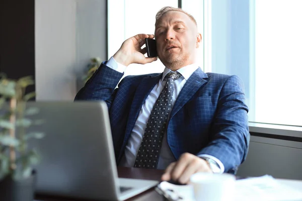 Middle aged businessman with laptop talking on telephone in modern office. — Stock Photo, Image