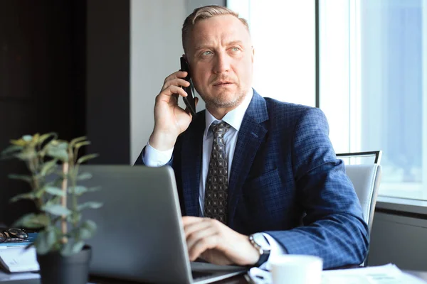 Středního věku podnikatel s notebookem mluvit na telefonu v moderní kanceláři. — Stock fotografie