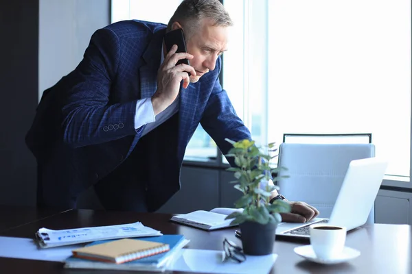 Бізнесмен середнього віку з ноутбуком розмовляє по телефону в сучасному офісі . — стокове фото