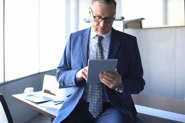 Серьезный зрелый финансовый консультант, сидящий за столом и проверяющий финансовый отчет с помощью цифрового планшета в офисе . — стоковое фото