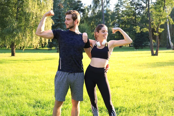 Alegre casal esportivo treinando juntos e mostrando bíceps no parque verde no dia ensolarado . — Fotografia de Stock