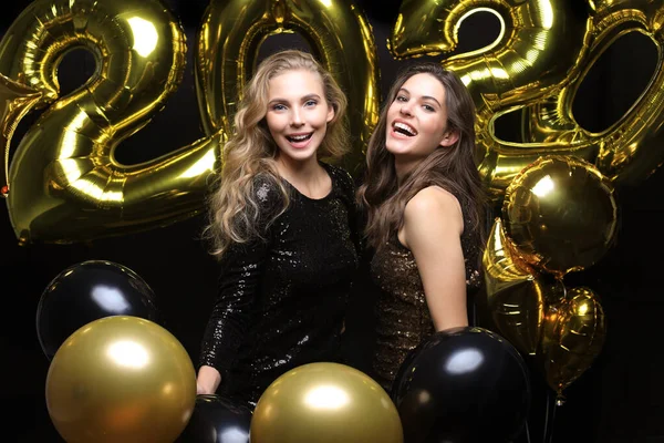 Two happy girls in shiny dresses posing while standing with gold colored 2020 number balloons on black background. — Stock Photo, Image