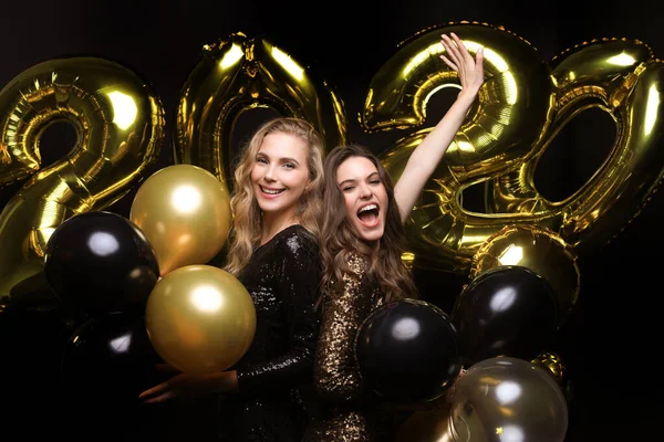 Duas meninas felizes em vestidos brilhantes posando enquanto de pé com ouro colorido 2020 balões número no fundo preto . — Fotografia de Stock