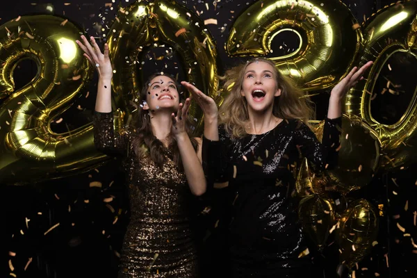 Duas meninas felizes em vestidos brilhantes posando enquanto de pé com ouro colorido 2020 balões número no fundo preto . — Fotografia de Stock