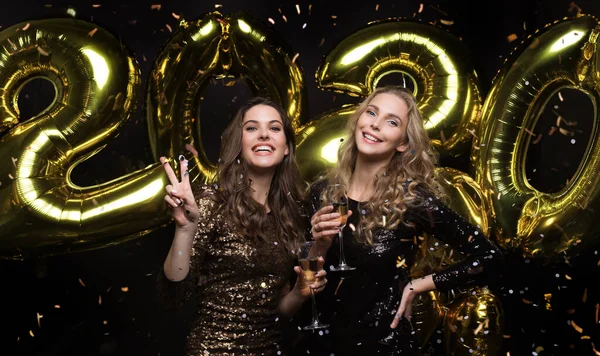 Duas meninas felizes em vestidos brilhantes posando enquanto de pé com ouro colorido 2020 balões número no fundo preto . — Fotografia de Stock