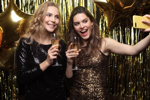 Beautiful girlfriends in high spirits take selfie at party on gold background. — Stock Photo, Image