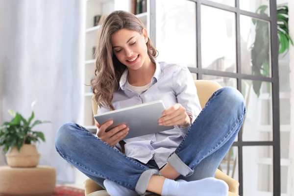 Mujer joven sonriente sentada en el sillón en la sala de estar y usando con su tableta . —  Fotos de Stock