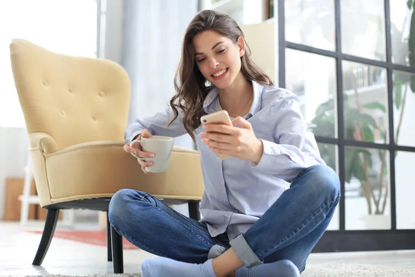 Mujer joven sonriente sentada en el suelo en la sala de estar y usando con su teléfono móvil . —  Fotos de Stock
