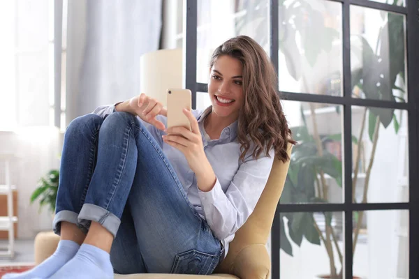 Mujer joven sonriente sentada en el sillón en la sala de estar y usando con su teléfono móvil . —  Fotos de Stock