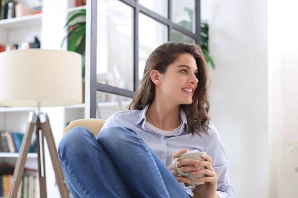 Glimlachend jong vrouwtje zittend in de fauteuil in de woonkamer, met een kopje koffie. — Stockfoto