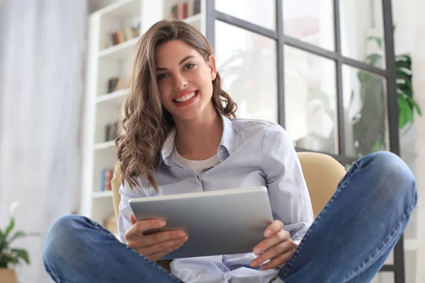 Mujer joven sonriente sentada en el sillón en la sala de estar y usando con su tableta . —  Fotos de Stock